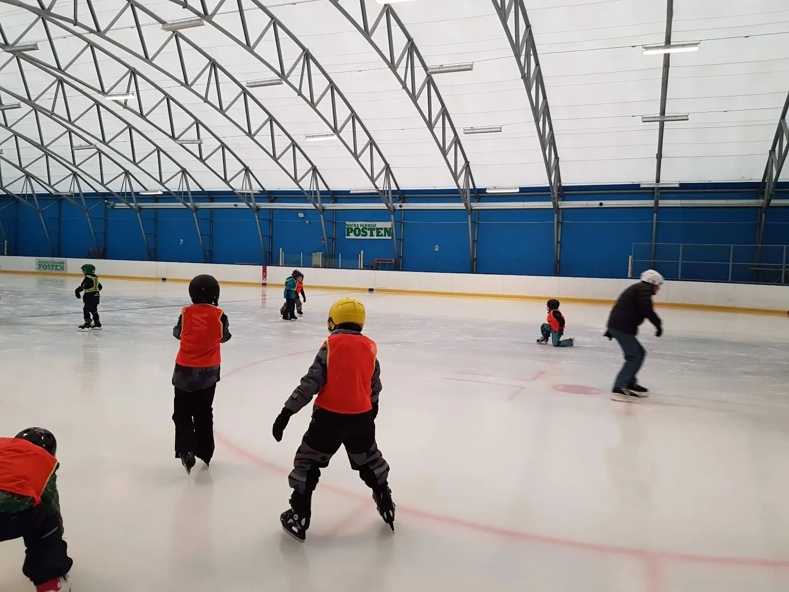 Young skaters and players on the ice inside the Nacka istält, a tent-like hall with a white arched roof, seen from the boards. — Nacka istält