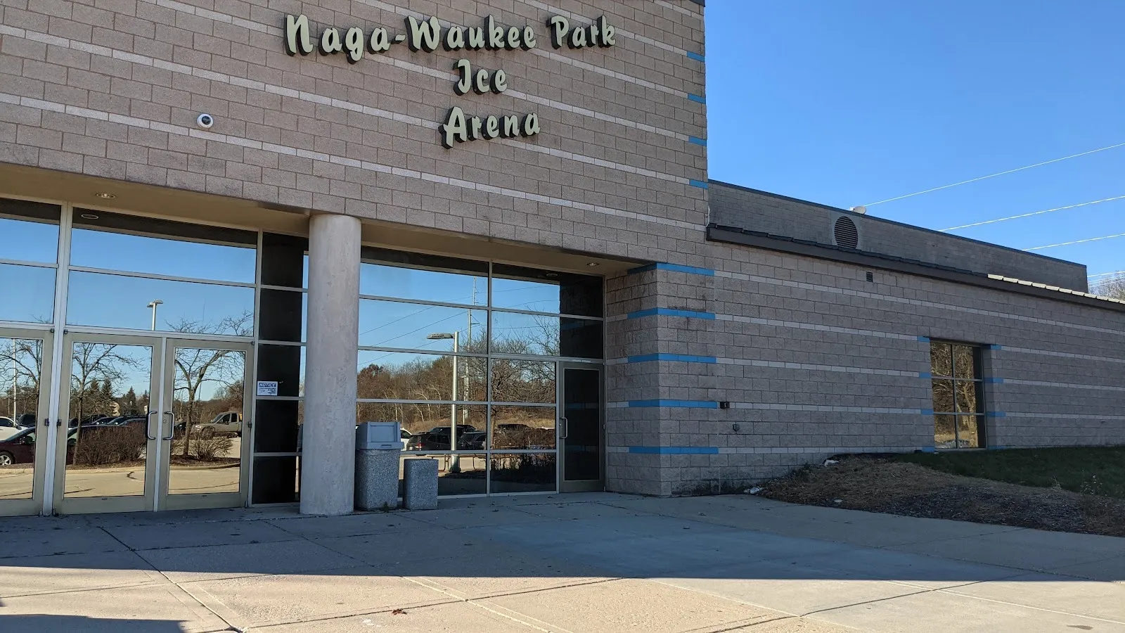 La entrada de Naga-Waukee Park Ice Arena, con su nombre en letra cursiva en el muro de bloque color canela sobre un vestíbulo y puertas de vidrio — Naga-Waukee Park Ice Arena