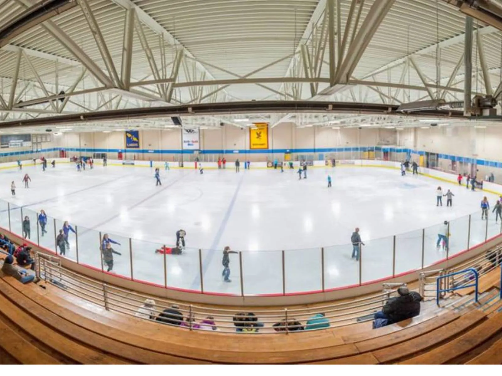 Amplia vista de ojo de pez desde las gradas de toda la pista de tamaño olímpico con patinadores recreativos en el hielo, gradas de madera en primer plano y mantas de equipos de hockey colgadas de las armaduras del techo. — Naga-Waukee Park Ice Arena