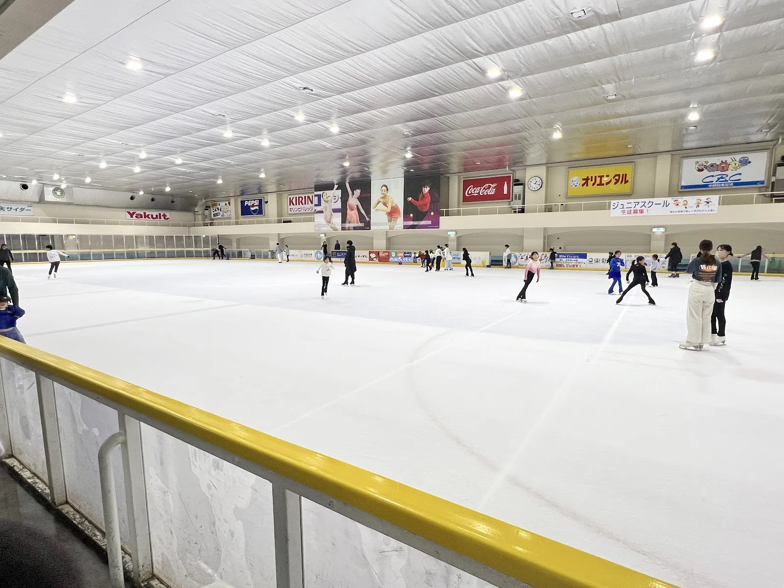 The ice sheet at Osu Skate Rink in the Nagoya Sports Center, seen from the yellow boards across the white ice, with sponsor signs along the far wall and a few skaters in the distance — Nagoya Sports Center (Osu Skate Rink)