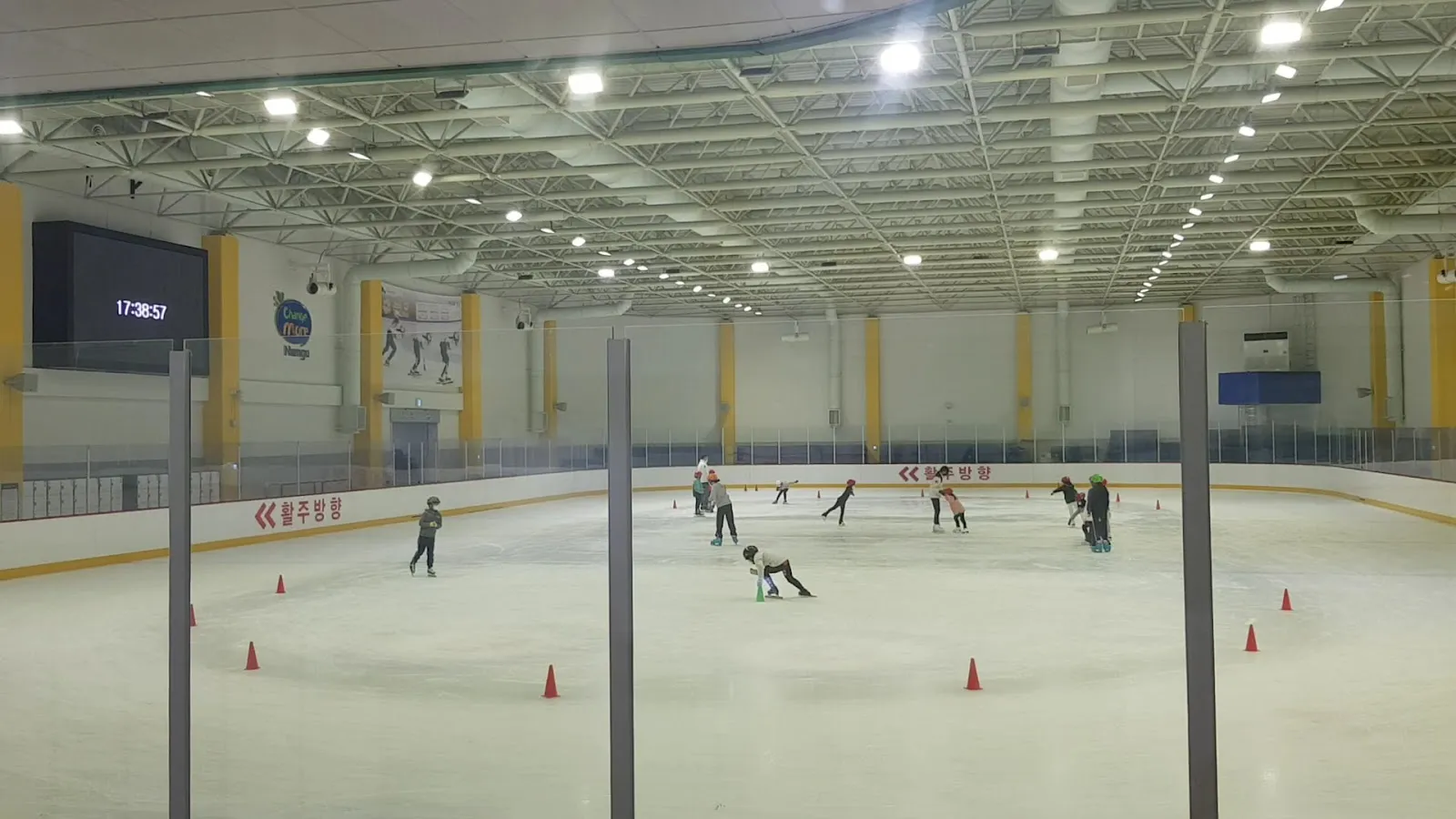 The wide ice sheet of Namgu Indoor Ice Rink with a few skaters far out on the ice under the steel-truss roof and orange cones. — Namgu Indoor Ice Rink