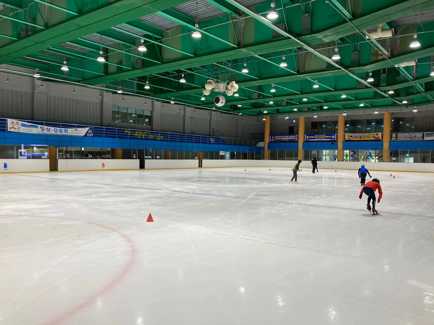 The ice rink of Namseon Park Gymnasium under its green steel roof, a wide view along the boards to the far stand with a few skaters and cones on the ice. — Namseon Park Gymnasium Ice Rink (Daejeon)