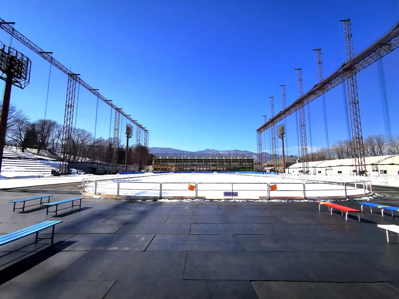 The oval's ice seen across the infield with benches along the edge, floodlight masts on both sides and snow at the track edge. — NAO ice OVAL (Chino International Skating Center)