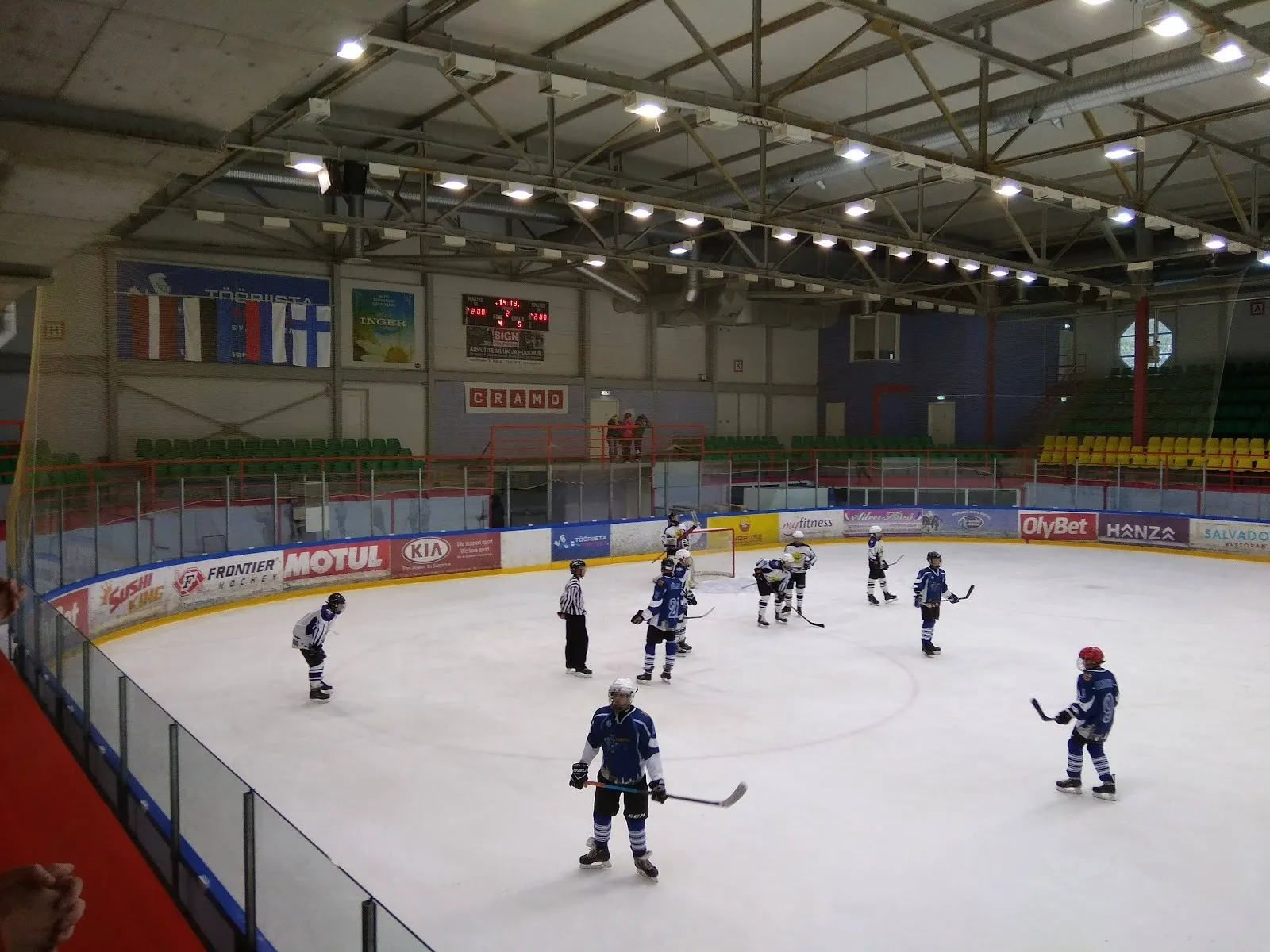Mängijad Narva jäähalli jääl, nende taga lipukaunistusega laudad, tribüünid ja teraskatus. – Narva jäähall