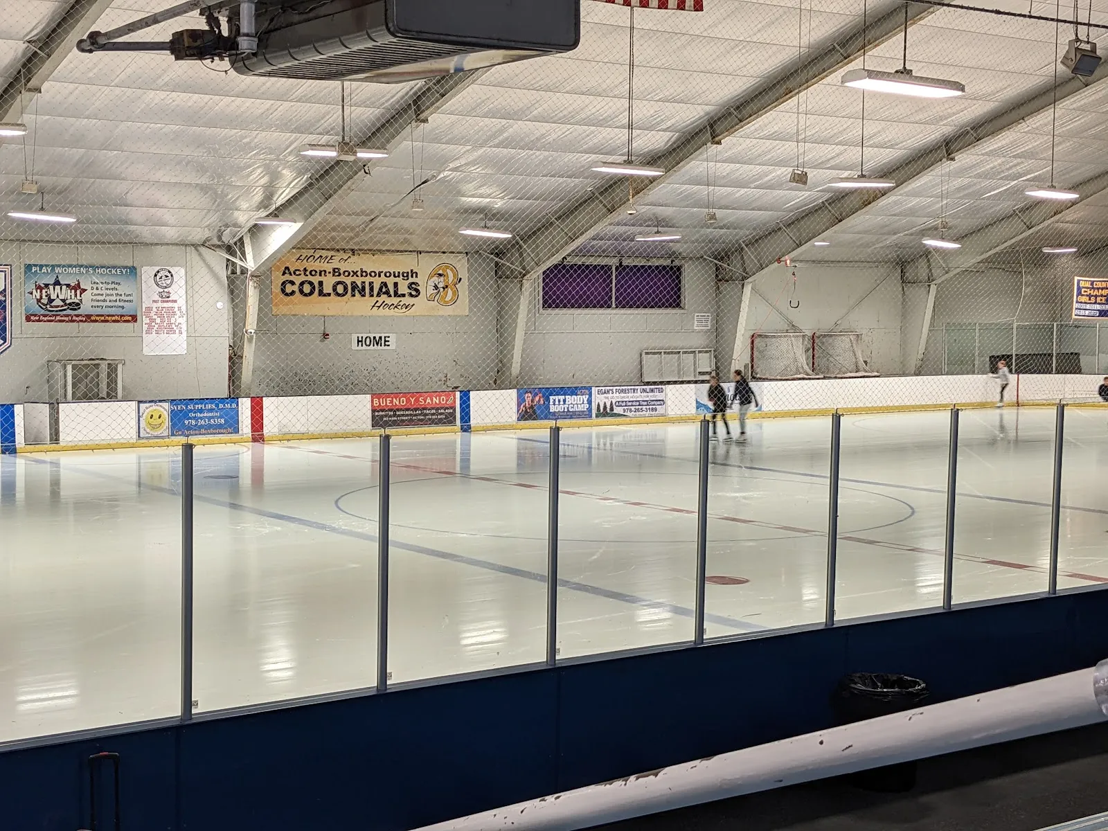 The ice seen through the glass, with an Acton-Boxborough Colonials banner on the far wall, sponsor boards along the dasher and a few skaters at the far end. — Nashoba Valley Olympia