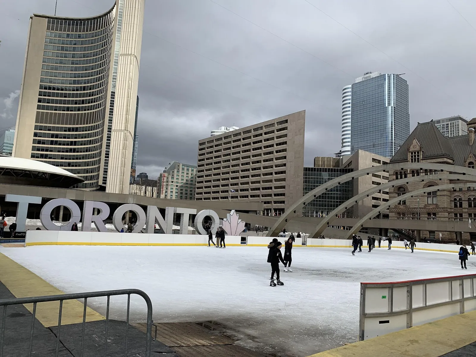 La pista de Nathan Phillips Square junto al gran letrero blanco de TORONTO, con unos cuantos patinadores en el hielo y la torre curva del Ayuntamiento y los arcos detrás, bajo un cielo gris. — Nathan Phillips Square Rink