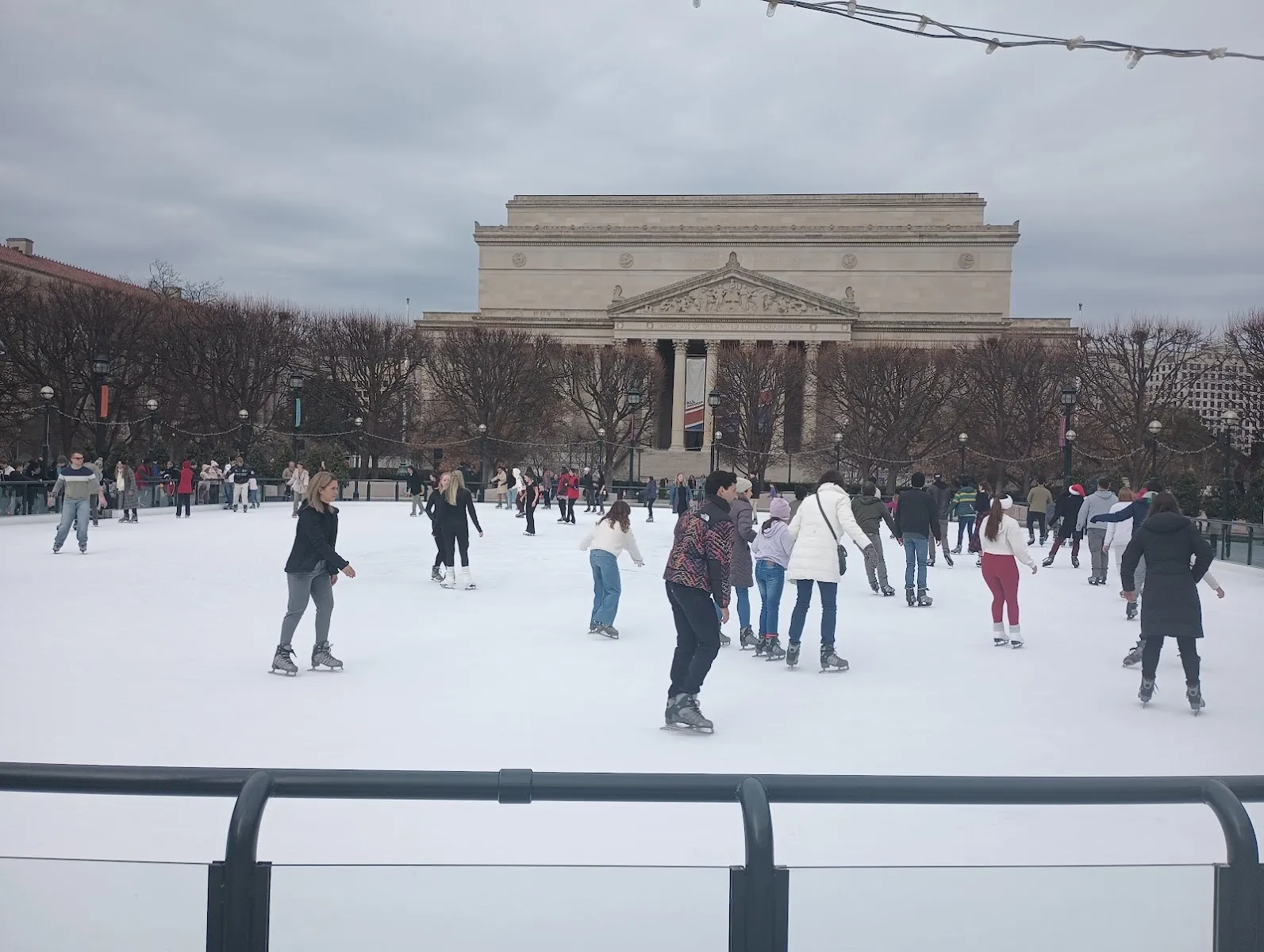 Skaters in the distance on the Sculpture Garden rink with the National Gallery's West Building rising behind the boards. — National Gallery of Art Sculpture Garden Ice Rink