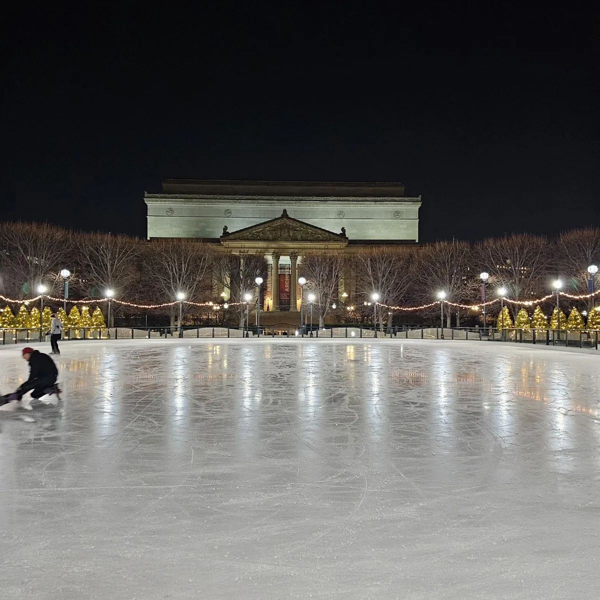 The Sculpture Garden rink at night, lit white with skaters far across the ice and the museum behind. — National Gallery of Art Sculpture Garden Ice Rink