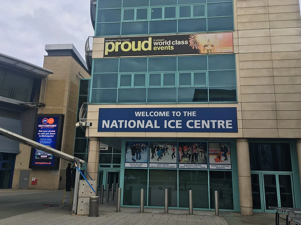 The main entrance of the National Ice Centre on Bolero Square: a tan-brick and glass facade under a blue 'Welcome to the National Ice Centre' sign, with photo panels of skaters in the doors. — National Ice Centre