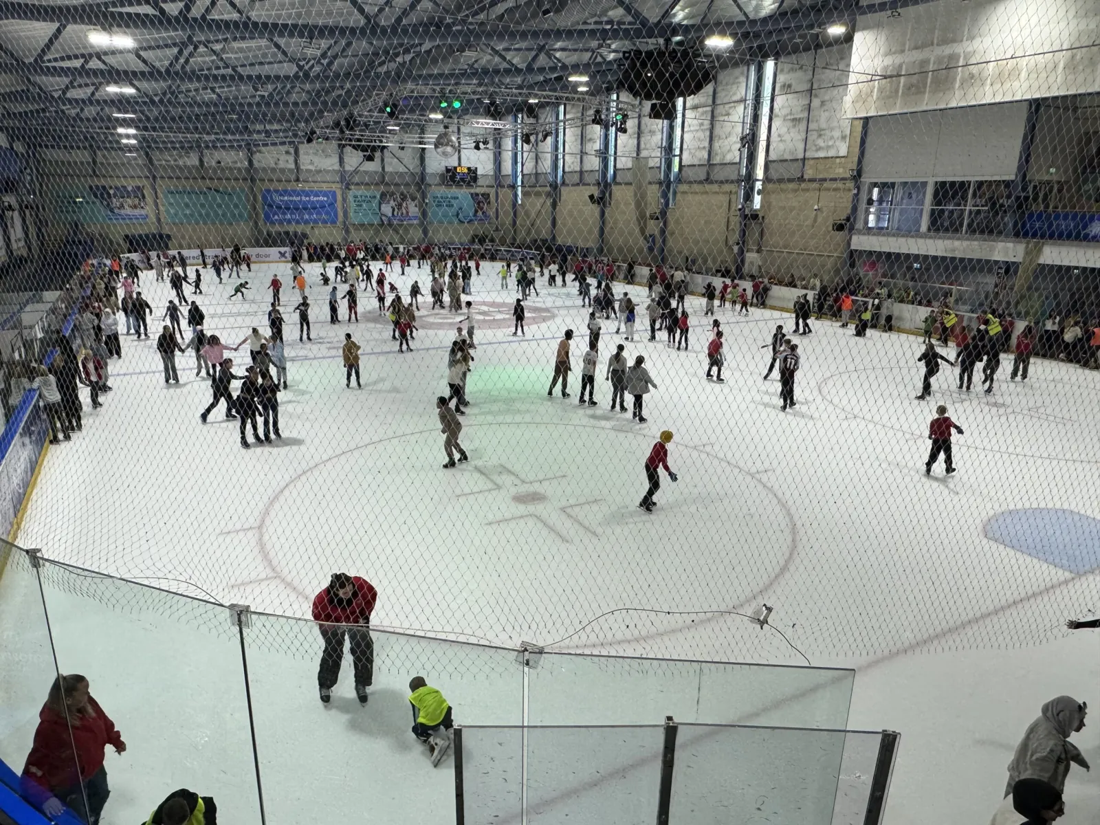 A wide view across the Nottingham rink during a public session, with skaters spread over the ice, the centre circle marked and a netted barrier along the side. — National Ice Centre
