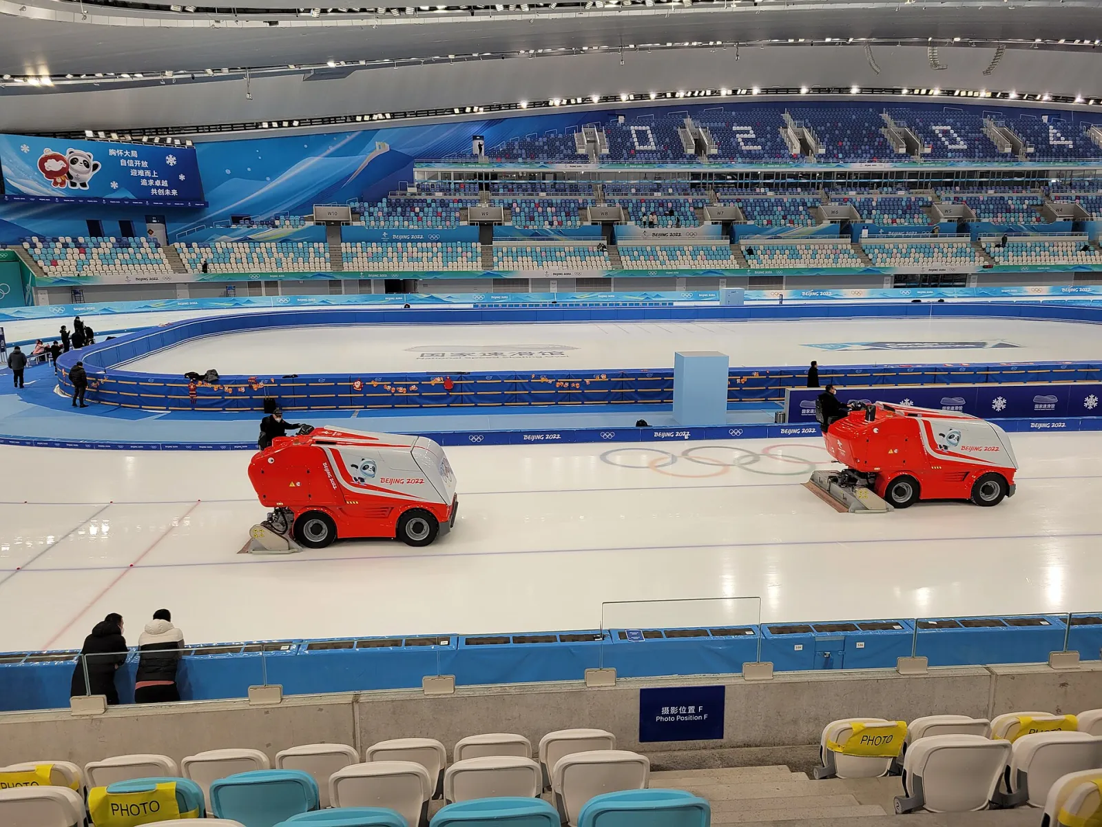 Two ice resurfacers on the oval's ice with the stands and the Olympic rings painted on the ice behind them — National Speed Skating Oval