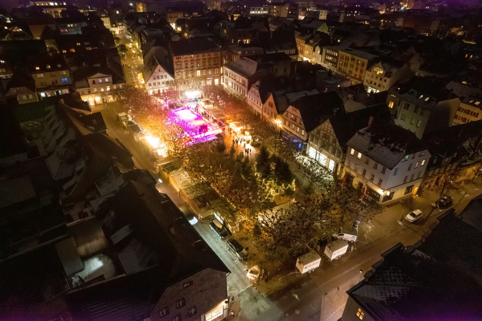 Night aerial view of the lit Holzminden Marktplatz, with the glowing natural-ice rink among the market huts under the trees. — Natur-Eisbahn Holzminden