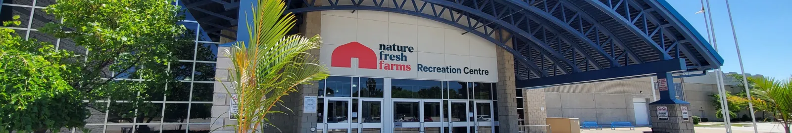 La entrada principal de vidrio del Nature Fresh Farms Recreation Centre con su letrero de logotipo rojo y blanco bajo una marquesina curva de acero azul, con palmeras en macetas junto a las puertas. — Nature Fresh Farms Recreation Centre