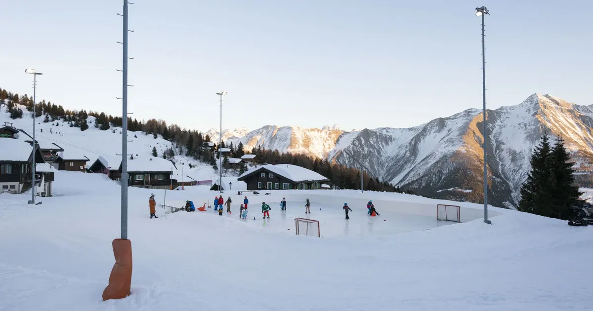 The boarded natural-ice rink at Bettmeralp with skaters and goals at the far end, floodlight masts and snowy peaks behind. — Natureisbahn Bettmeralp