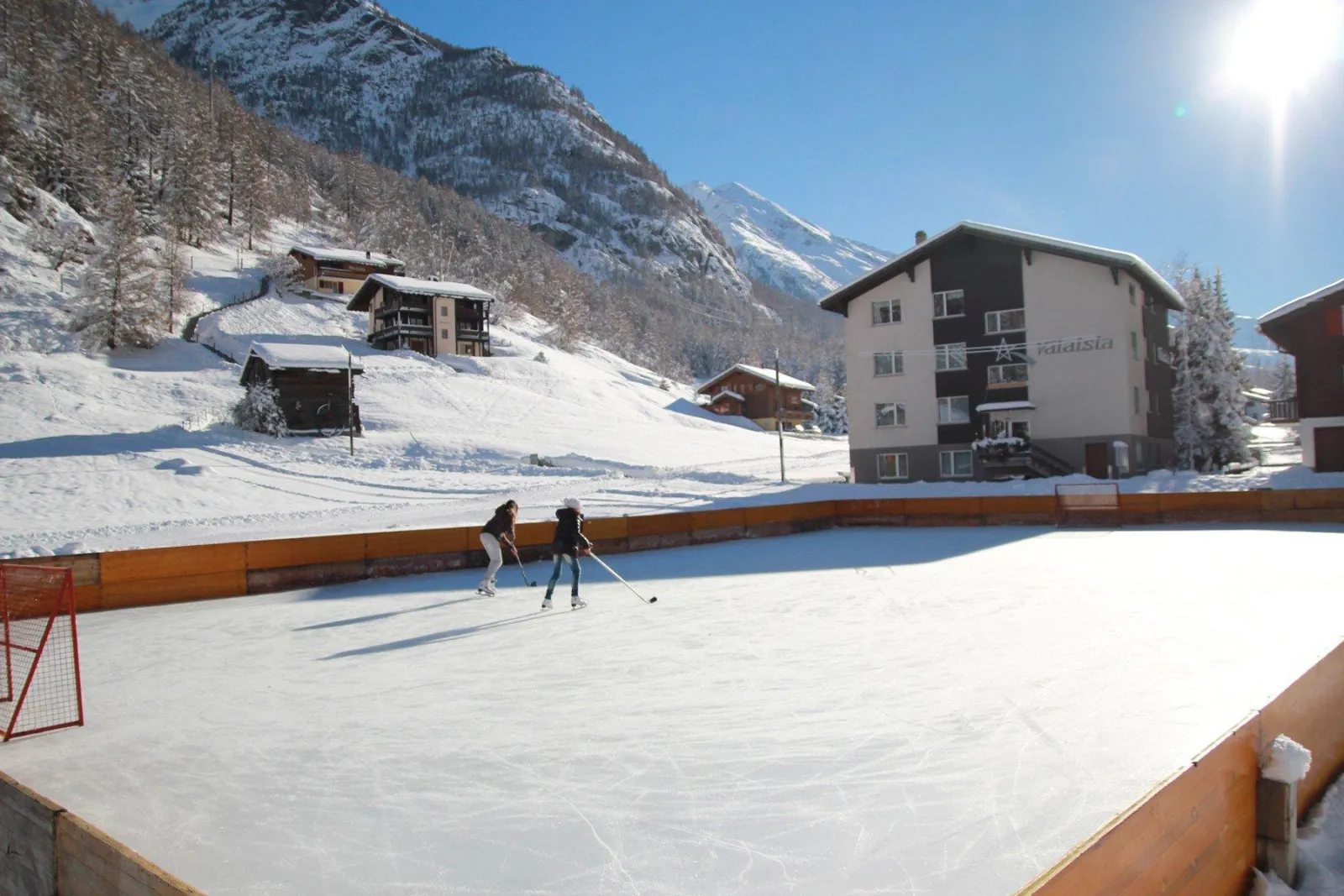 The boarded natural-ice rink in Randa with two skaters at the far end, chalets on the slope and snowy peaks behind. — Natureisbahn Randa