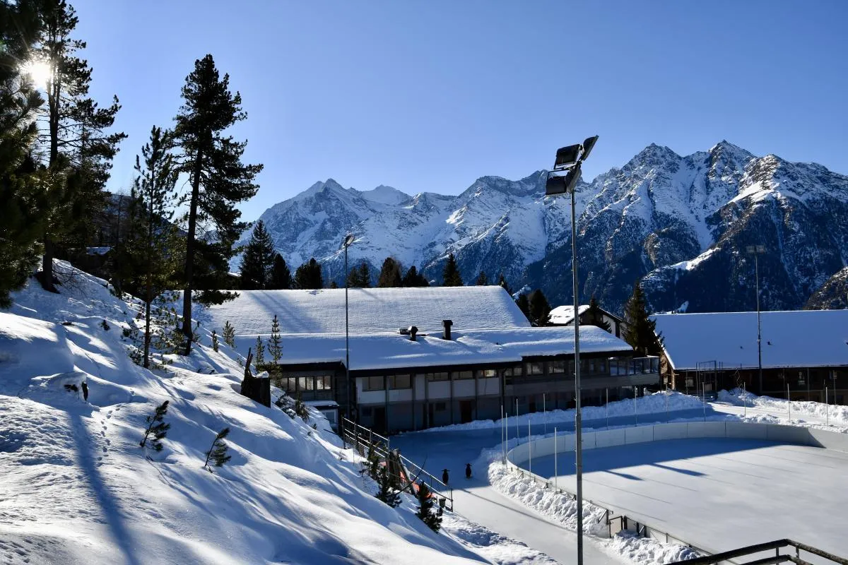 The Sportzentrum Grächen seen from the slope above, with the rink beside the building and the Alps behind. — Natureisbahn Sportzentrum Grächen
