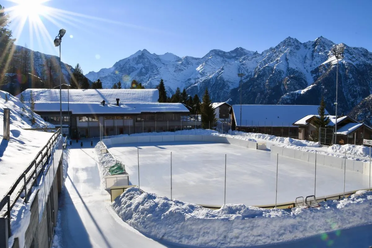 The boarded natural-ice rink at the Sportzentrum Grächen in winter sun, with the sports centre and snow-covered mountains behind. — Natureisbahn Sportzentrum Grächen