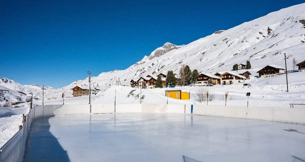 The natural ice rink at Juppa in Avers on a sunny winter day, rink boards along the ice with chalets and snowy slopes behind – Natureisfeld Avers-Juppa