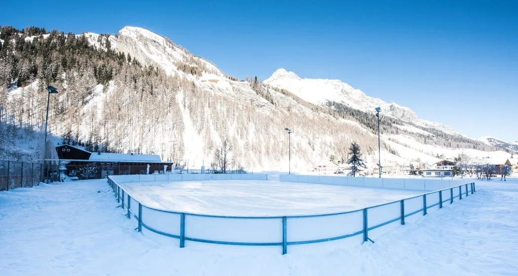 The natural ice field at Camping Splügen, a blue-fenced ice surface in the snow below the mountains — Natureisfeld Camping Splügen