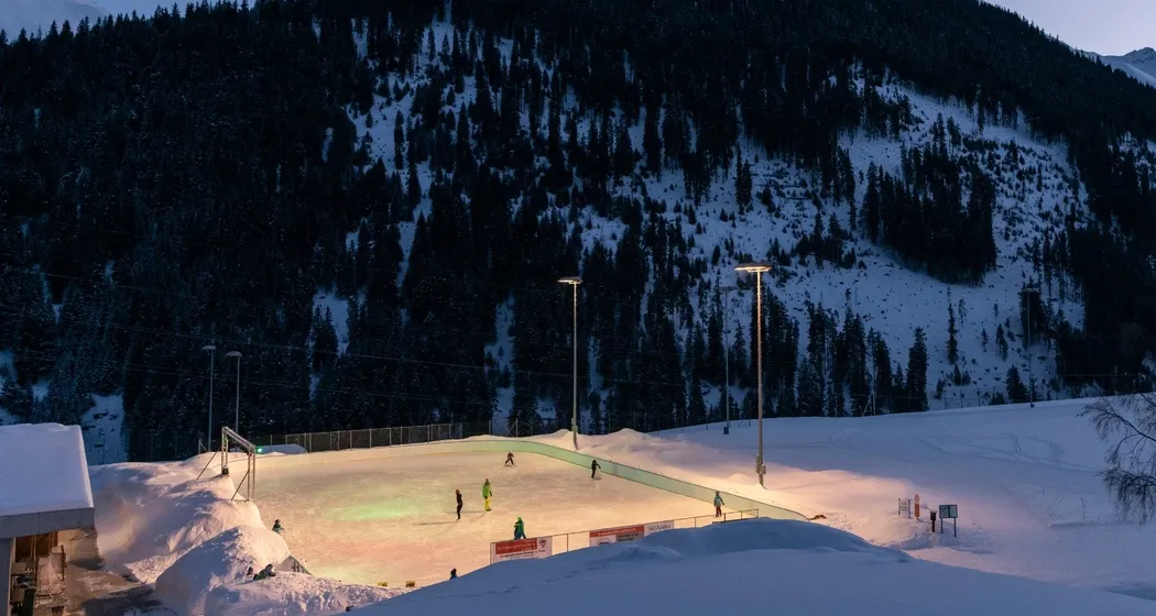 The Sedrun natural-ice field seen from the slope above in the evening, lit by floodlights, with a forested valley side behind. — Natureisfeld Sedrun