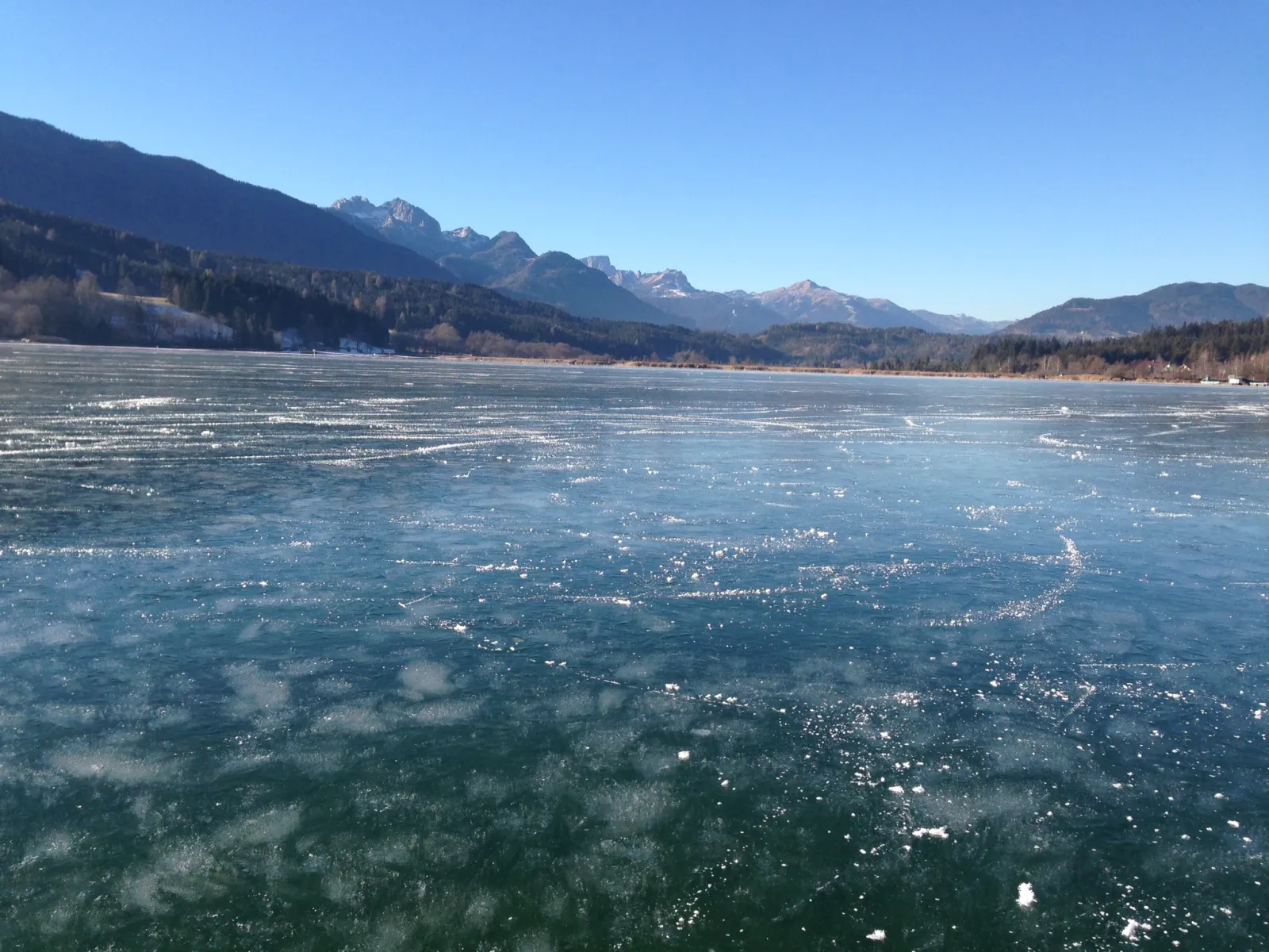 The frozen surface of the Pressegger See scored by skate tracks, with forested slopes and mountains along the far shore under a clear sky. — Natureisfläche Pressegger See