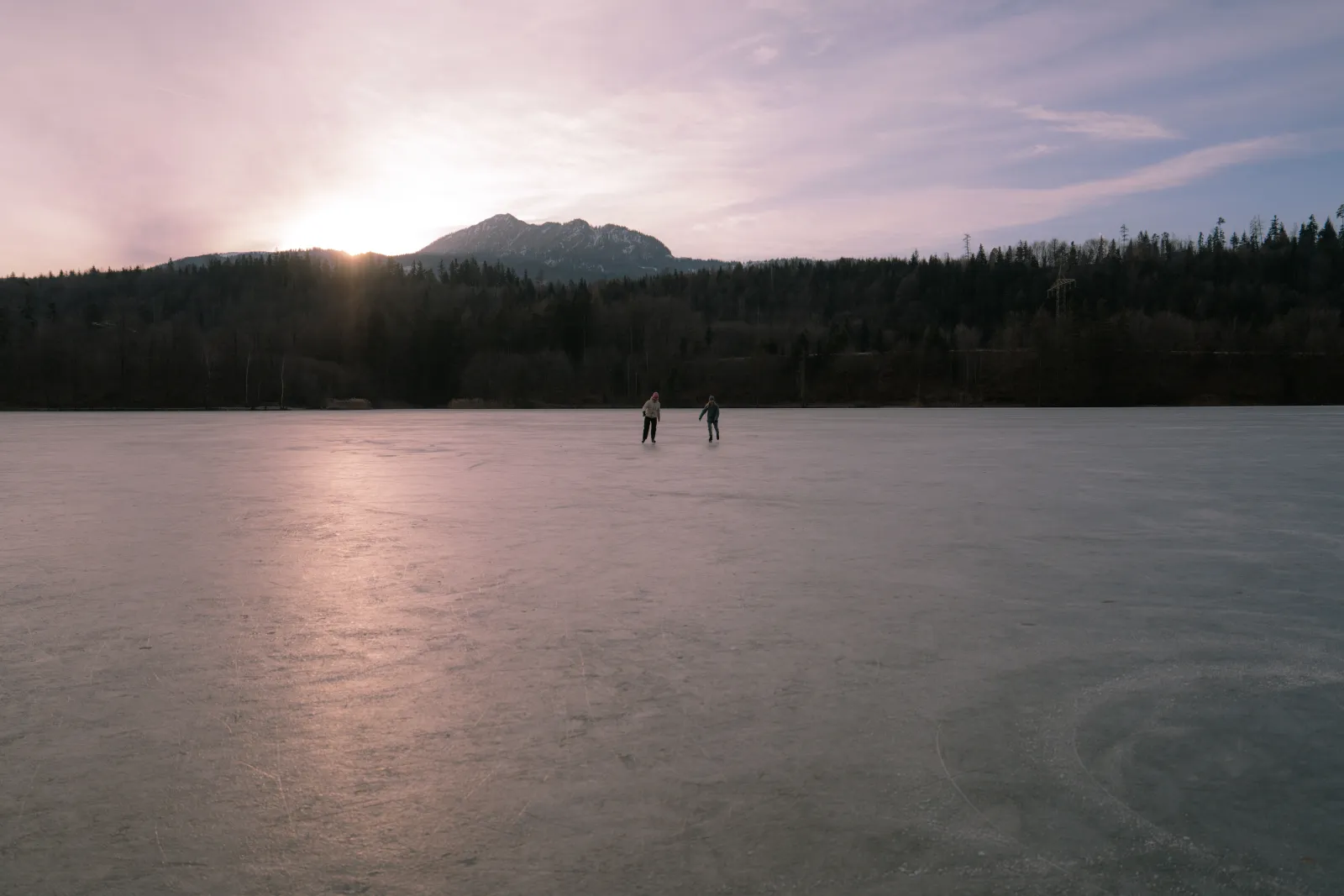 Two skaters far out on the frozen Reintaler See at dusk, with a pink sky over a dark wooded shoreline and a mountain beyond. — Natureisfläche Reintaler See