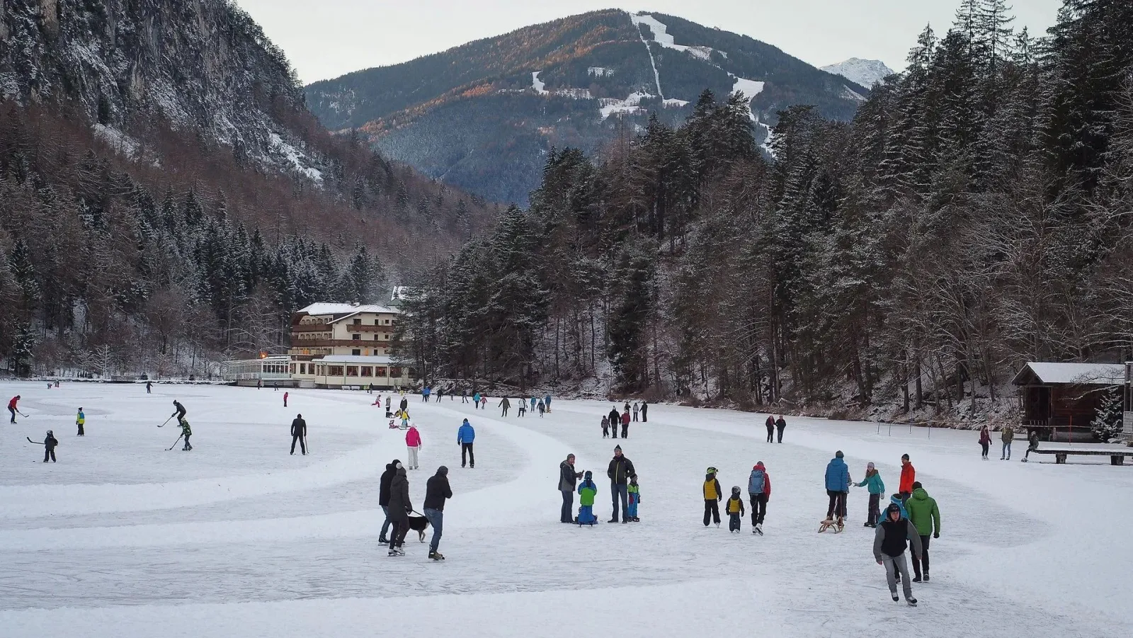 Skaters spread across the frozen Tristacher See, with a lakeside building and a wooded mountain valley behind. — Natureisfläche Tristacher See