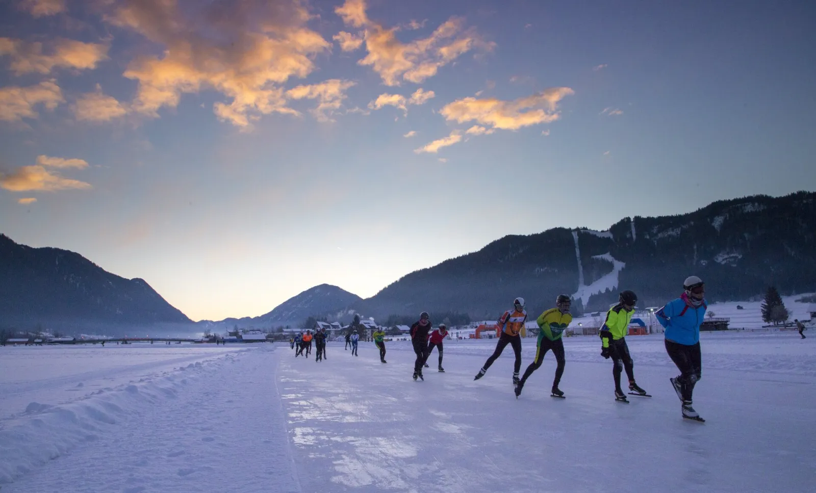 Skaters strung out across the frozen Weissensee at dusk, under a pink and orange sky, with snow-covered mountains along the shore. — Natureisfläche Weissensee