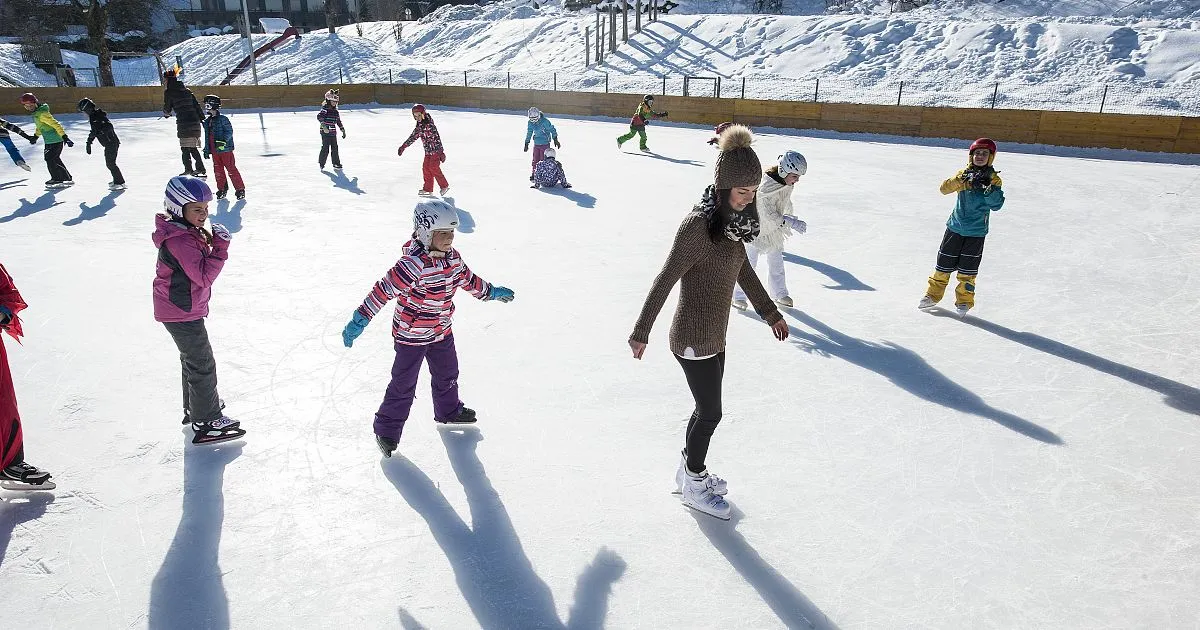 Children and adults skating on the Altenmarkt rink in bright winter sun, with long shadows across the ice. — Natureislaufplatz Gemeindepark Altenmarkt