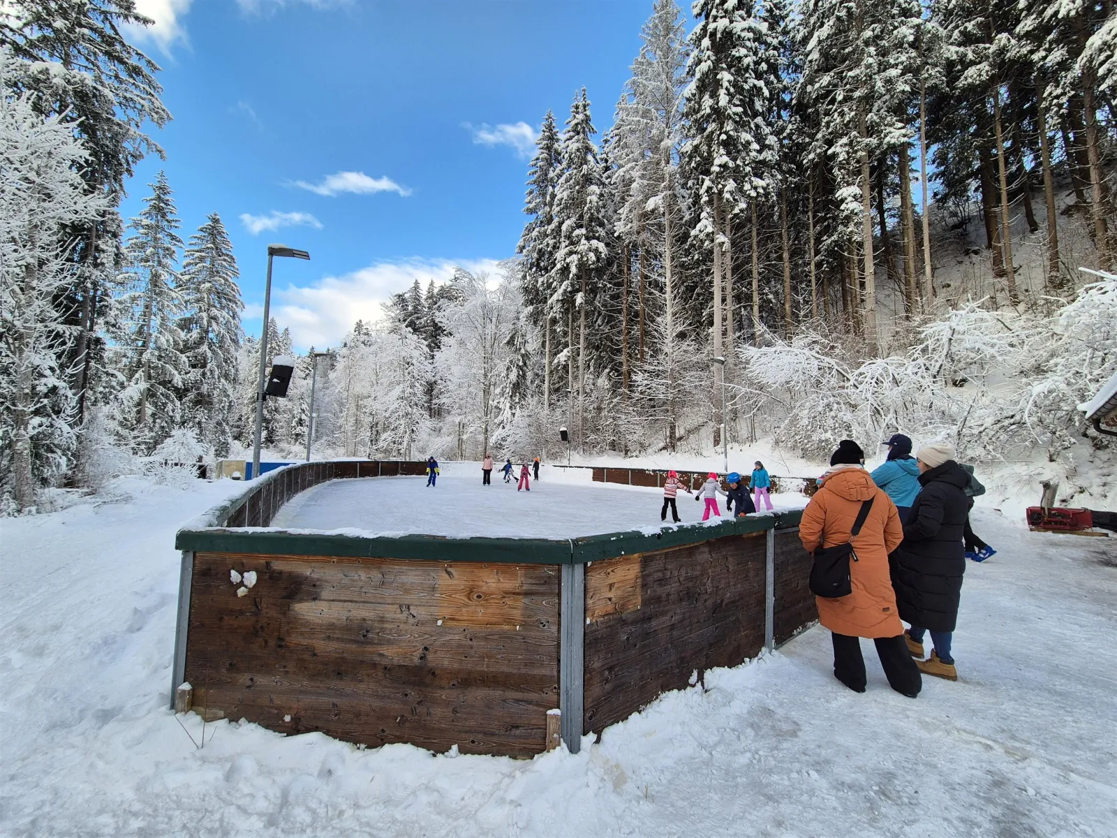 The boarded natural-ice rink at Going in a snowy spruce forest, with skaters at the far end and visitors along the wooden boards. — Natur-Eislaufplatz Going