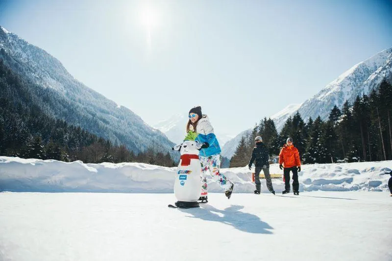 Skaters spread across the sunny natural-ice rink in the Stubai valley, with snow-covered mountains behind. — Natureislaufplatz Klaus Äuele