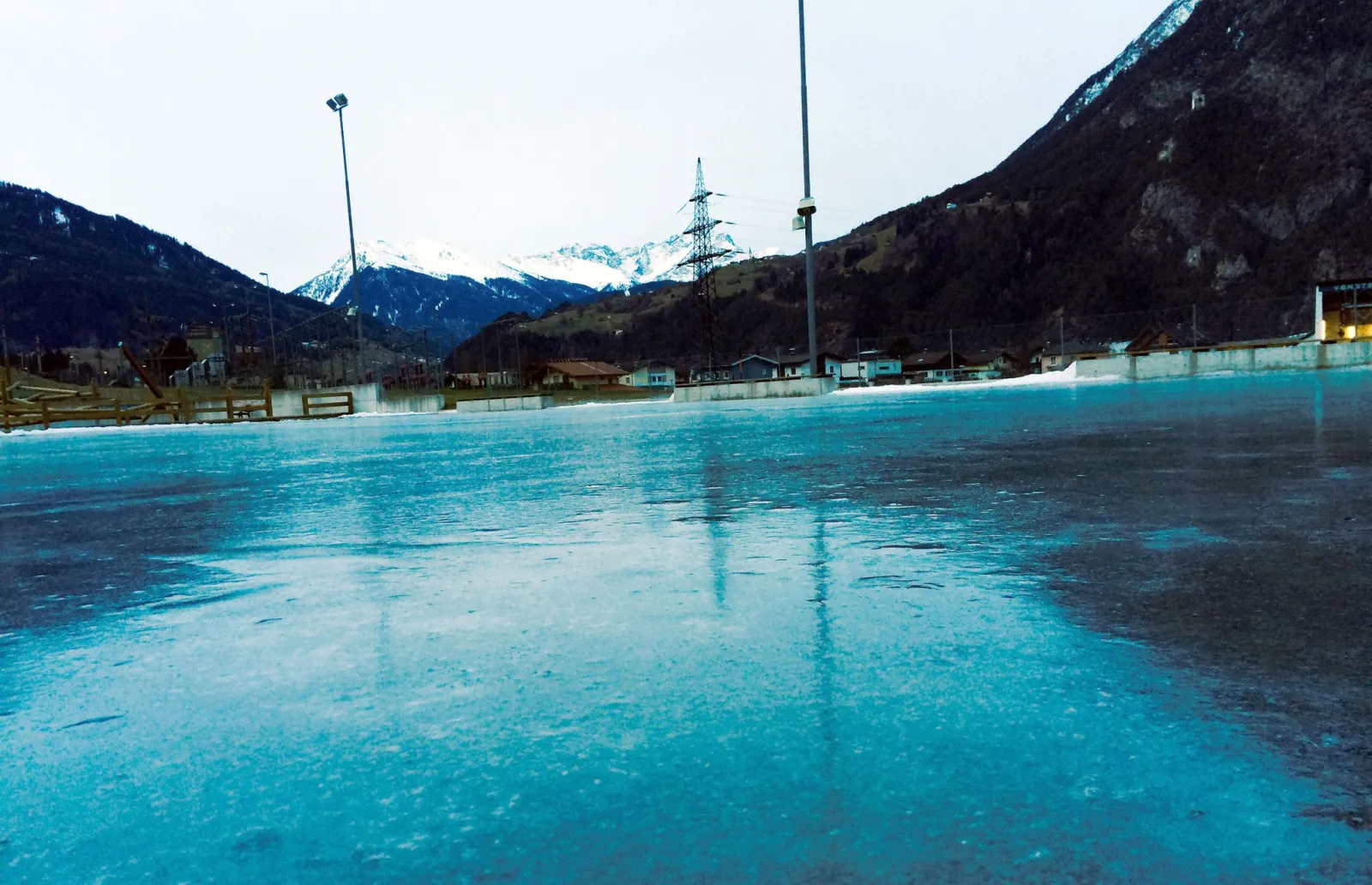 The turquoise ice of the Eislaufplatz Riefe in Zams, with floodlight masts and mountains on both sides of the valley. — Eislaufplatz Riefe