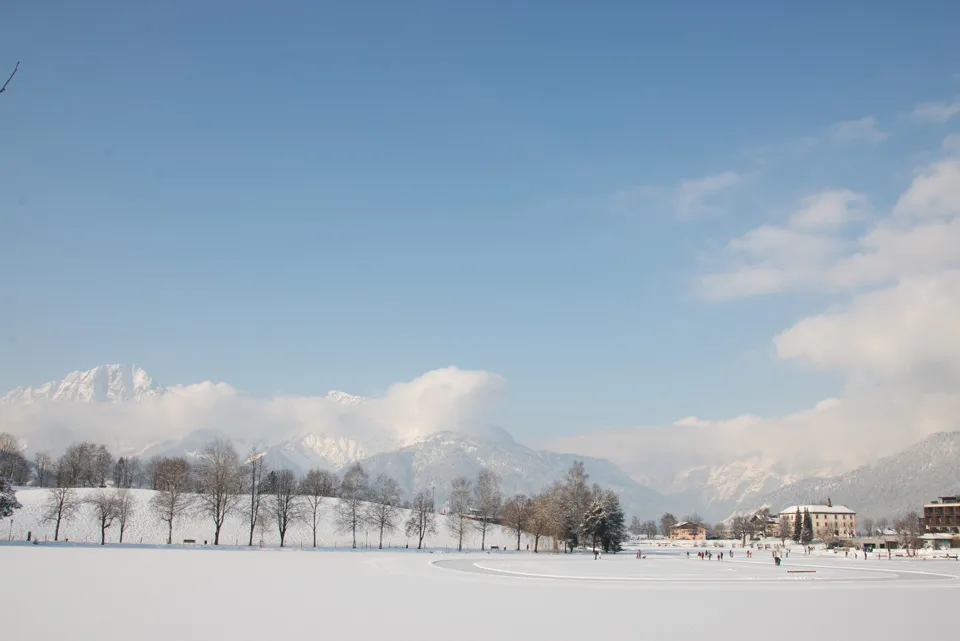 The snow-covered Ritzensee in Saalfelden with a row of bare trees along the shore and mountains on the horizon. — Natureislaufplatz Ritzensee
