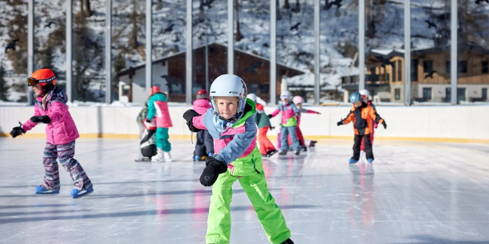 Children in colourful snowsuits skating together on the Sölden rink, with a glazed wall and alpine chalets behind the boards. — Eislaufplatz Sölden