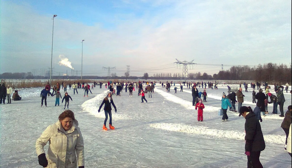 Een grote menigte schaatsers op de open natuurijsbaan van Oosterhout in de winterzon, met lichtmasten en hoogspanningsmasten aan de horizon. – Natuurijsbaan De Elskens