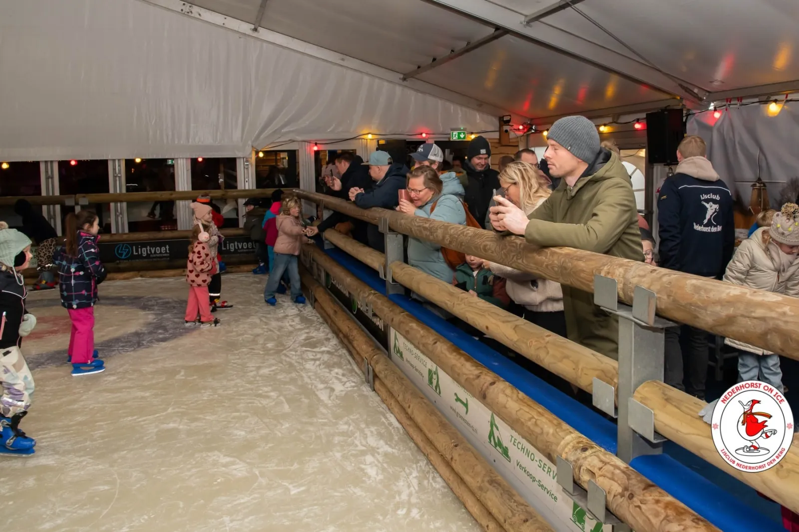 Kinderen schaatsen op afstand op het ijs in de overdekte tent van Nederhorst on Ice, met rechts de houten boarding en toeschouwers die eroverheen leunen onder de lichtsnoeren. – Nederhorst on Ice (Nederhorst den Berg)
