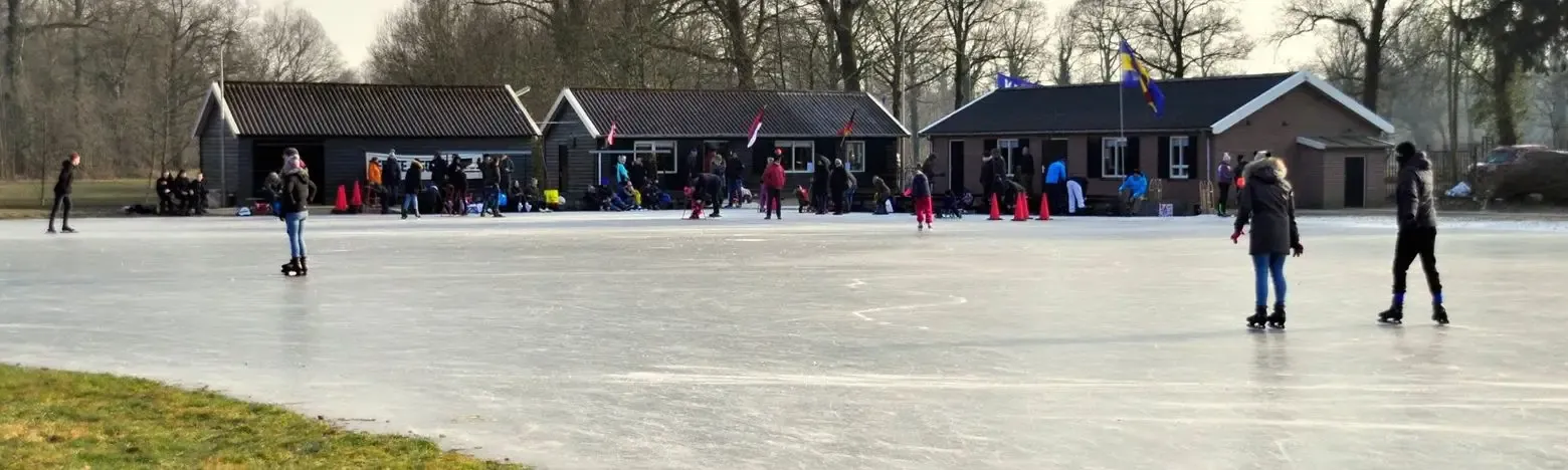 The low dark clubhouse buildings of the Needse IJsclub with their flags behind the swept ice, with a few skaters and visitors in front and bare trees behind. — IJsbaan De Kooweide (Needse IJsclub, Neede)
