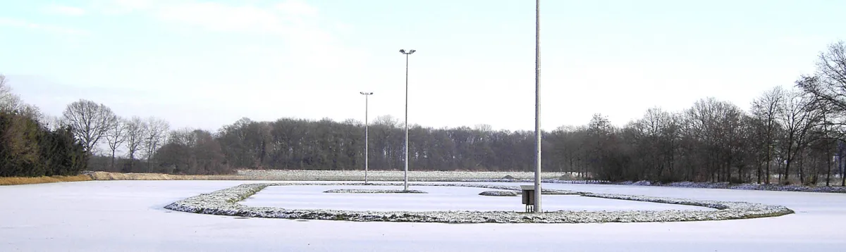 The empty frozen oval of IJsbaan De Kooweide at Neede under a pale winter sky, with a ring of rime along the inner edge, tall floodlight masts and bare trees around it. — IJsbaan De Kooweide (Needse IJsclub, Neede)