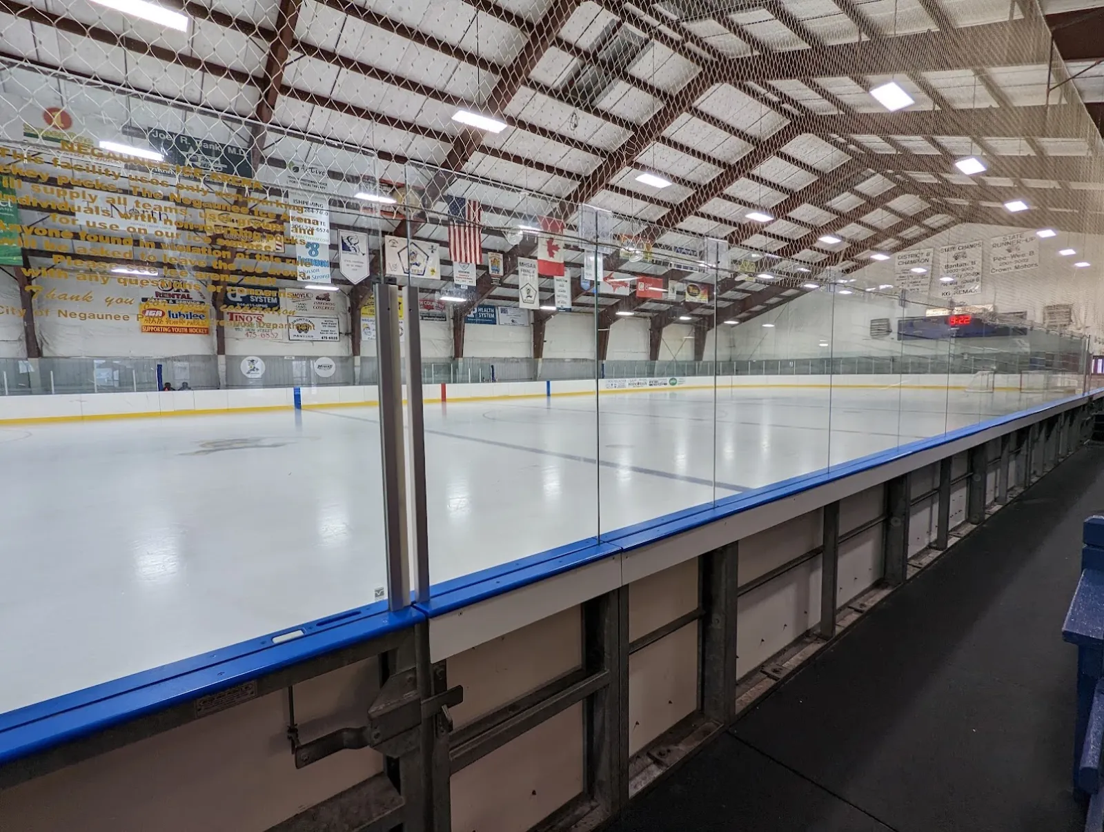 An empty sheet of ice from the glass at the boards, with banners and flags hanging from the steel roof trusses. — Negaunee Ice Arena