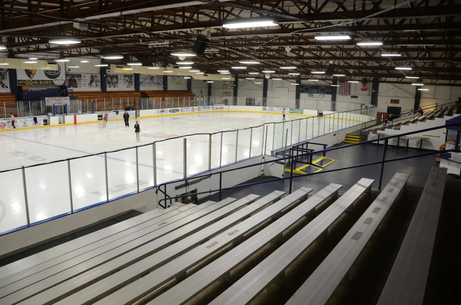 Vista elevada desde las gradas de toda la pista del Nelson Center, con vallas y cristal alrededor de la pista, mantas de patrocinadores y una manta de "Let's Go Jr. Blues!" en la pared del fondo, y algunos patinadores en el hielo — Nelson Center Ice Rink