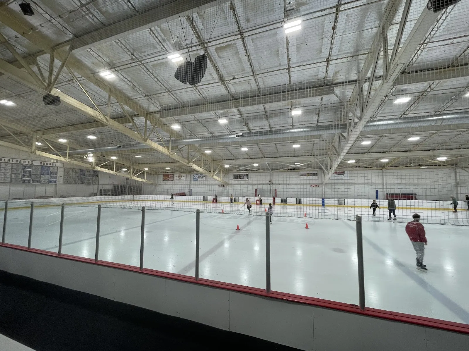 Wide interior view of the ice sheet from rink-side, with a handful of distant skaters and a coach working around traffic cones, a "RENEGADES HOCKEY" wall sign, and HOME/VISITORS boards visible under the roof trusses. — Nelson Sports Complex (Rolling Meadows)