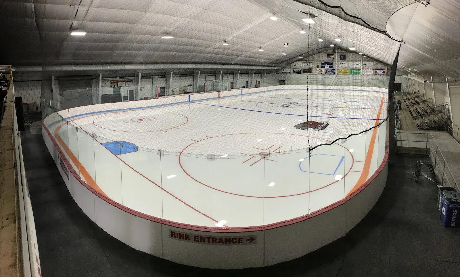 Una amplia vista de la pista vacía dentro de la arena cerrada, con vallas de borde naranja, un letrero de "Rink Entrance" en las vallas cercanas y gradas a lo largo del lado opuesto. — Nelson Withington Skating Facility