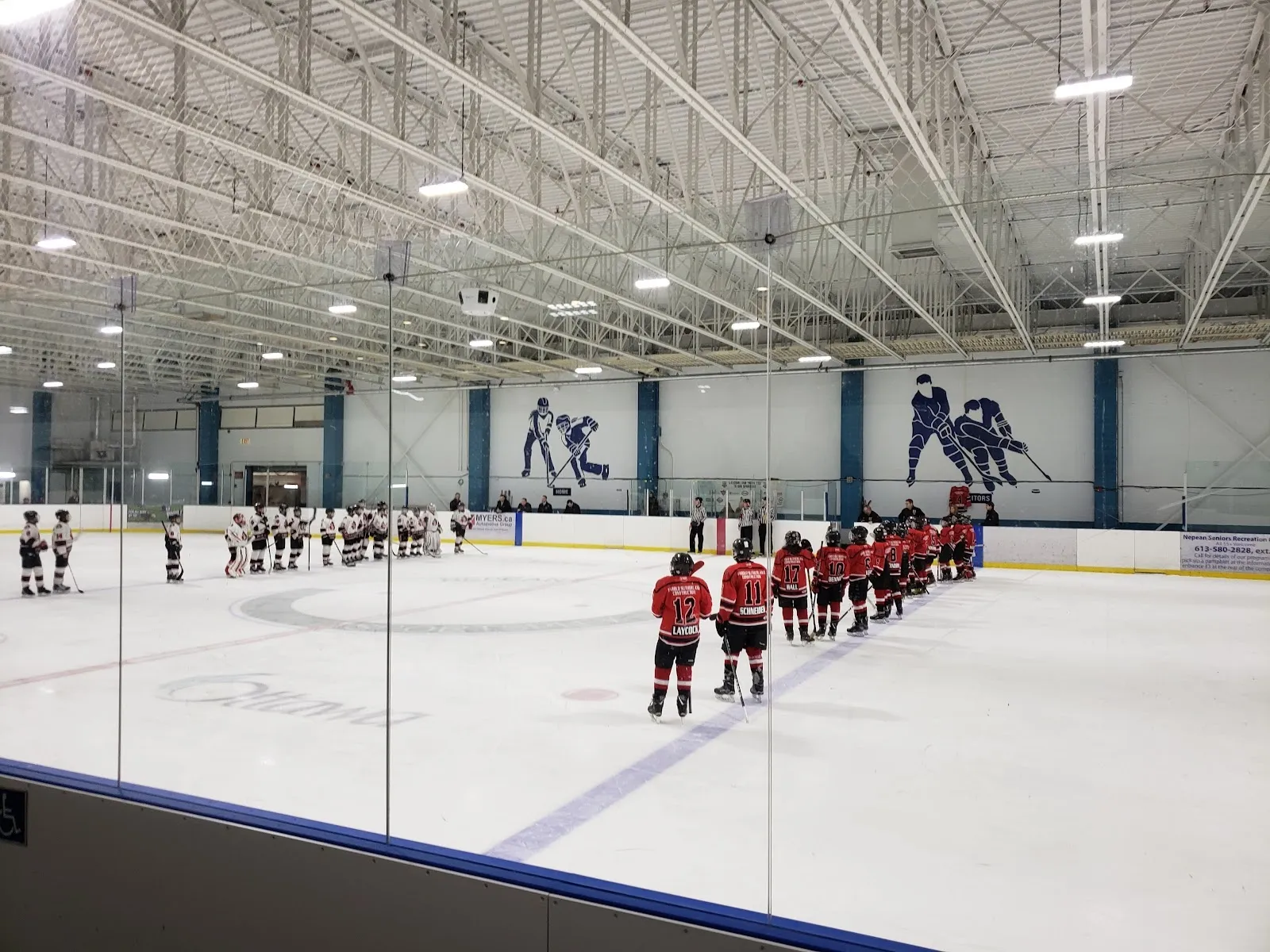 A team lined up on the blue line facing a white-uniformed team across a wide sheet, a blue-and-white wall and a high steel roof behind them. — Nepean Sportsplex