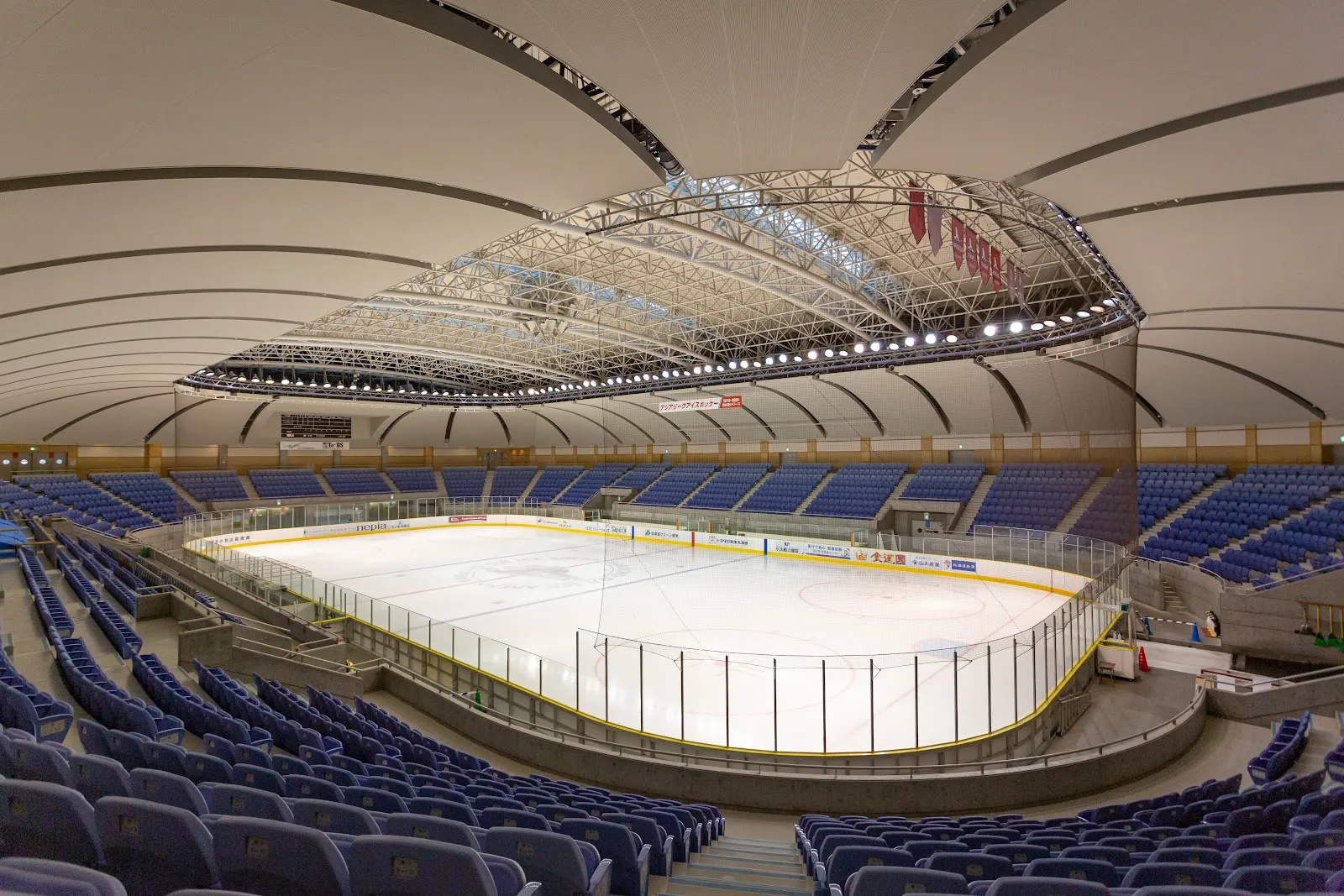 The empty ice sheet of the nepia Ice Arena in Tomakomai seen from the stands, with rows of blue seats and the sweeping white roof over the rink — nepia Ice Arena (Hakucho Arena)