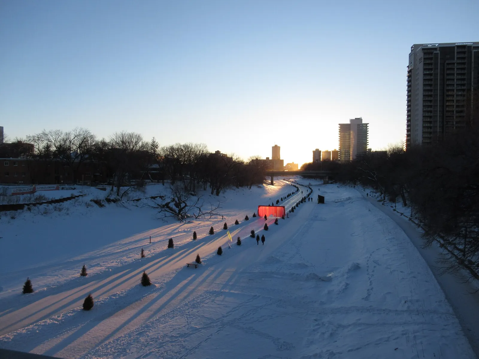 El Nestaweya River Trail sobre el río Red congelado al atardecer: un carril de patinaje despejado con algunos patinadores y señales del sendero, y edificios de la ciudad más allá — Nestaweya River Trail at The Forks