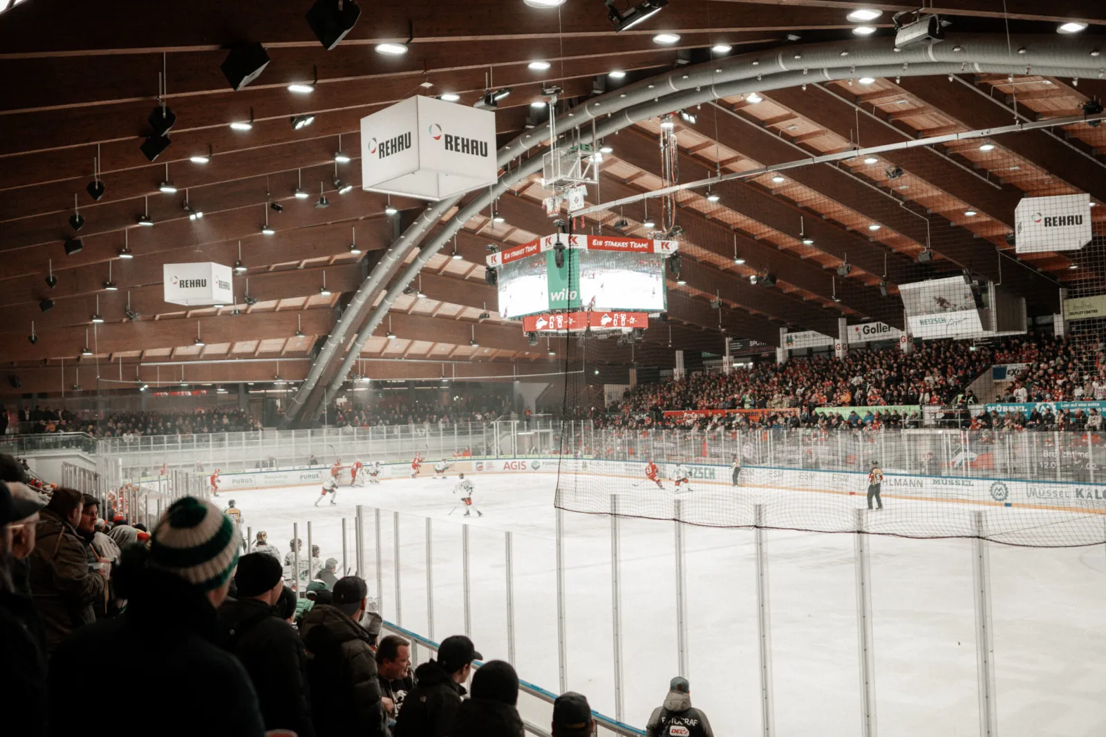 Ein DEL2-Heimspiel in der NETZSCH-Arena, von der Tribüne aus gesehen: die ganze Eisfläche, die geschwungenen Holzbinder des Dachs, der Videowürfel und eine volle Tribüne. – NETZSCH-Arena Selb