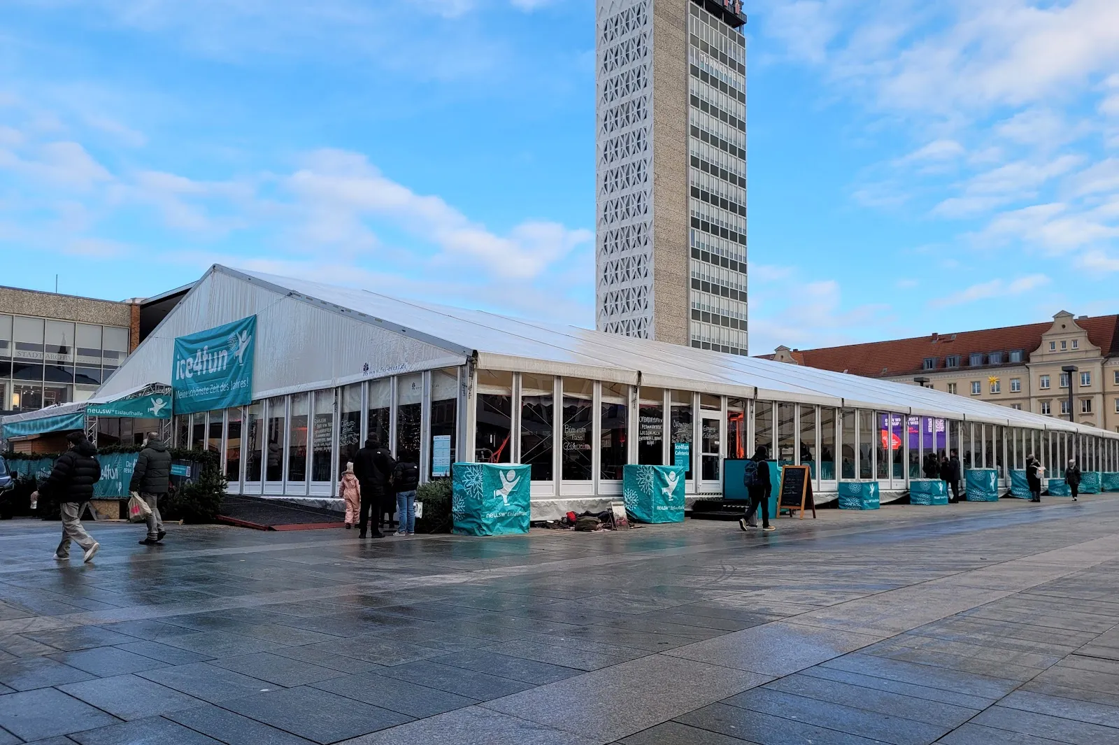 The long glazed skating-hall tent on the Neubrandenburg market square with the tower block of the old town hall behind. — neu.sw Eislaufhalle ice4fun