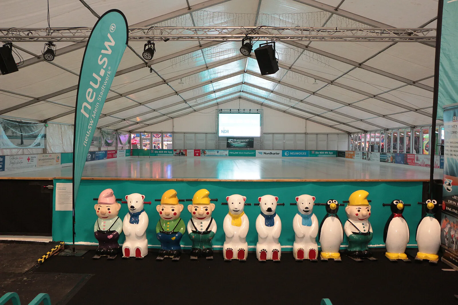 The heated skating hall under its white tent roof, with the empty ice and a row of penguin and bear skating aids along the boards. — neu.sw Eislaufhalle ice4fun