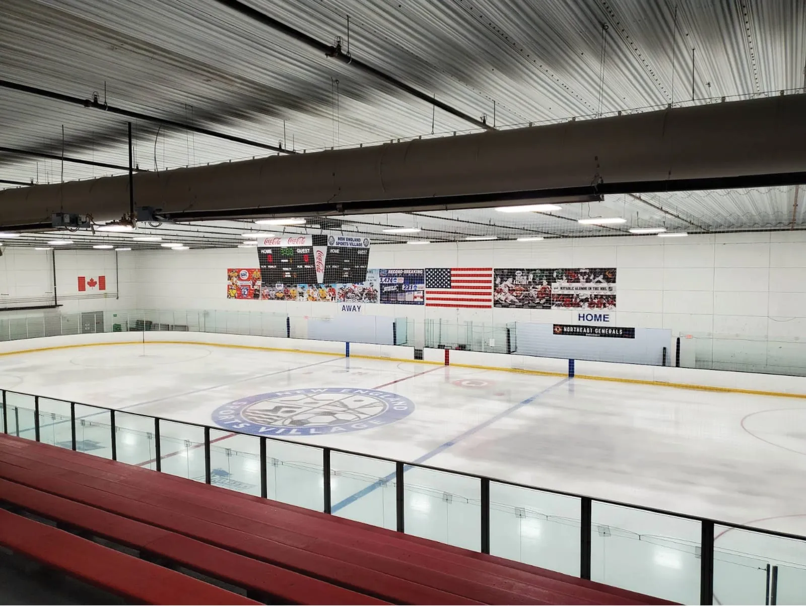 Una de las pistas NHL desde las gradas rojas, con el logotipo de NESV en el centro de la pista y banderas y marcadores en la pared del fondo. — New England Sports Village