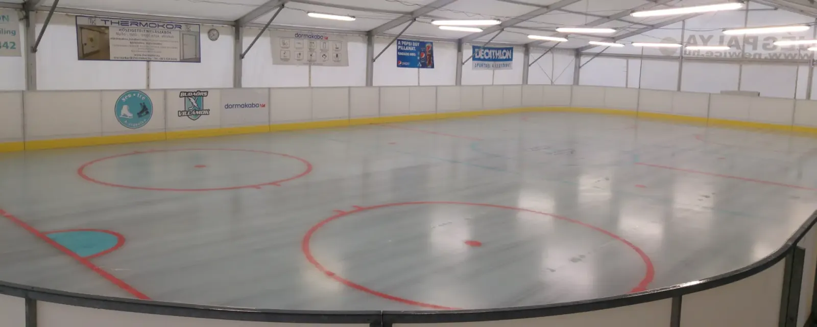 Üres jég a sátorral fedett New Ice jégpályán, piros bedobókörökkel, sárga palánkszegéllyel és a fehér sátortetővel – New Ice Jégpálya